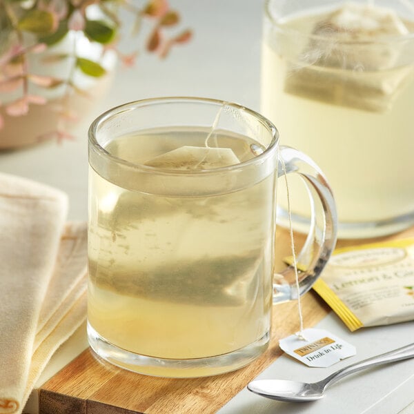 A glass mug of Twinings Lemon & Ginger tea with a tea bag in it on a wooden board.