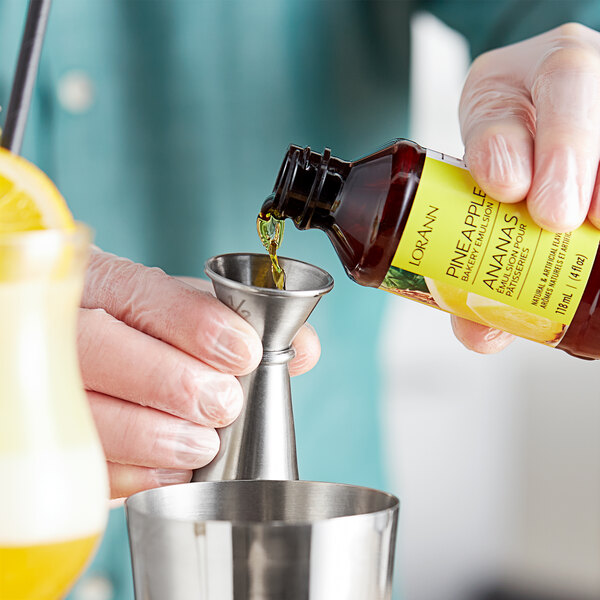 A hand pouring LorAnn Oils Pineapple Bakery Emulsion from a bottle into a metal cup.