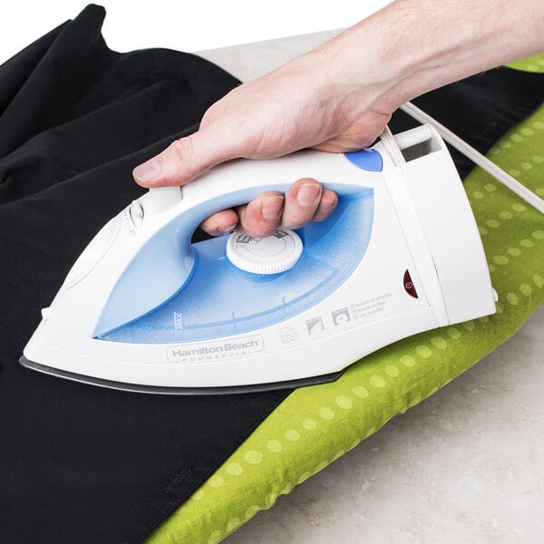 A person using a Hamilton Beach white and blue iron to iron black and green fabric on an ironing board.