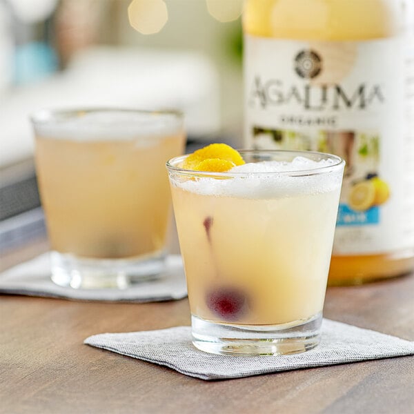 Two glasses of Agalima sweet and sour mix drinks with fruit on top on a table.