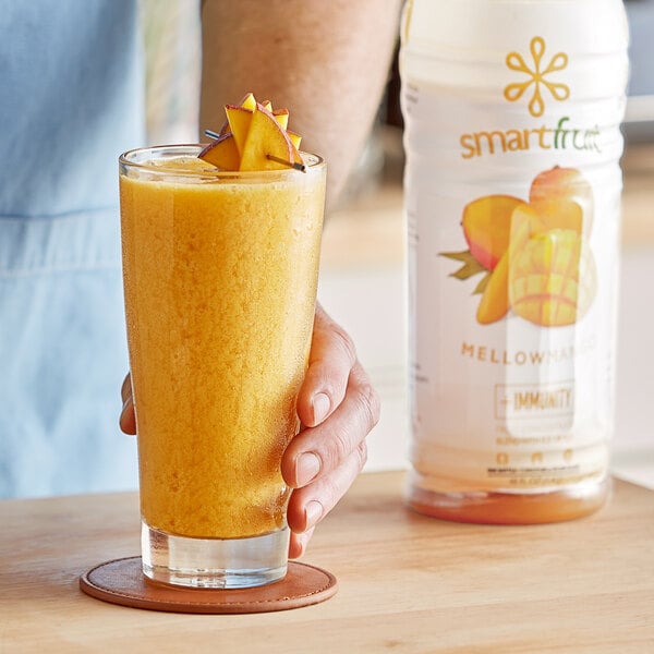 A hand holding a glass of yellow Smartfruit Mellow Mango puree beverage with fruit on top.