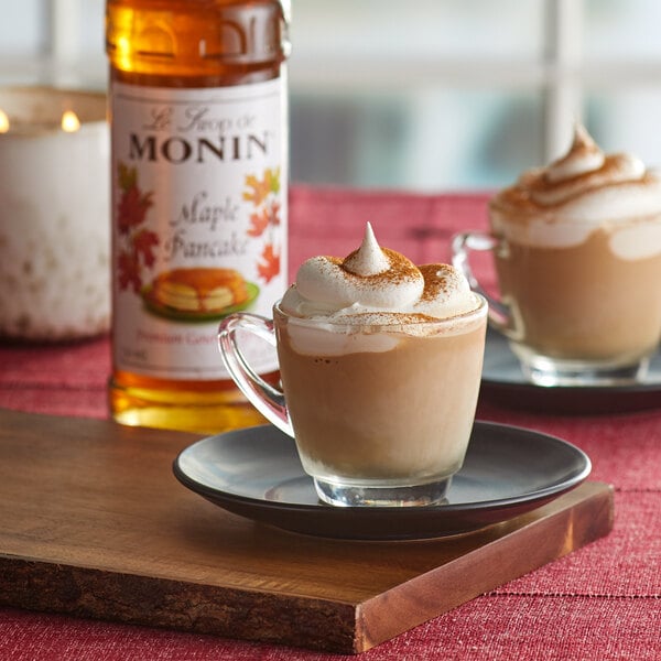 A glass cup of coffee with whipped cream and a bottle of Monin Premium Maple Pancake flavoring syrup.