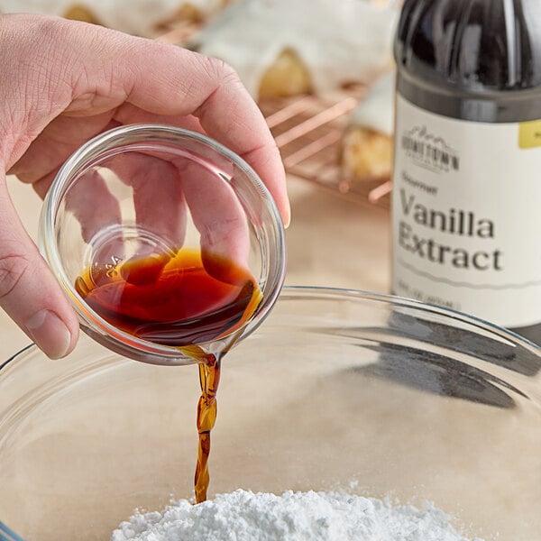 A hand pouring brown liquid from a Hometown Provisions Gourmet Pure Vanilla Extract container into a glass.