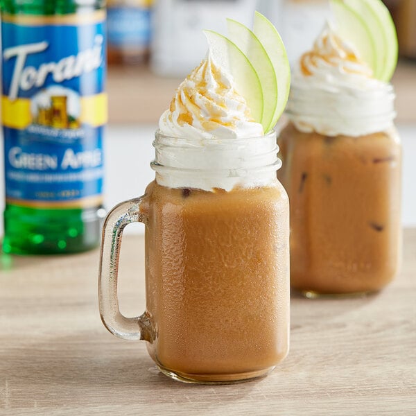 A glass mug of brown liquid with whipped cream and caramel with a bottle of Torani Sugar-Free Green Apple Flavoring Syrup in the background.