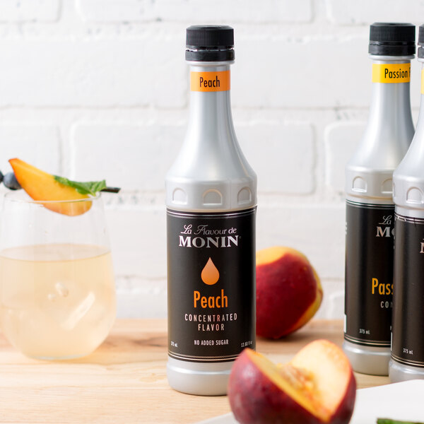 A white case of 3 Monin Peach Concentrated Flavor bottles on a table with a glass of peach flavored liquid.