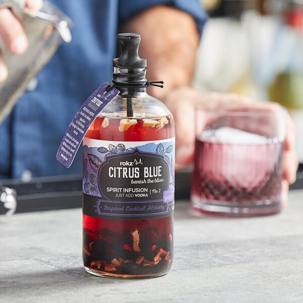 A bottle of Rokz Citrus Blueberry Cocktail Infusion being poured into a glass with a black straw.