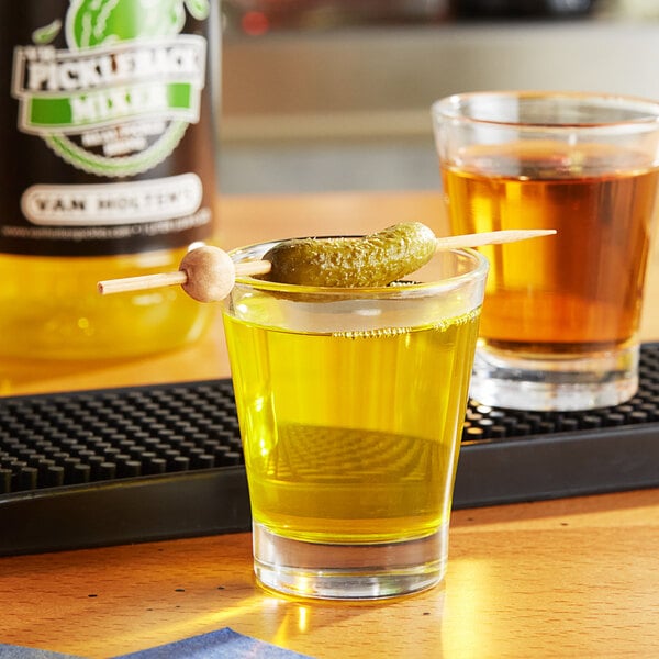 A glass of yellow liquid with a toothpick holding a pickle over it.