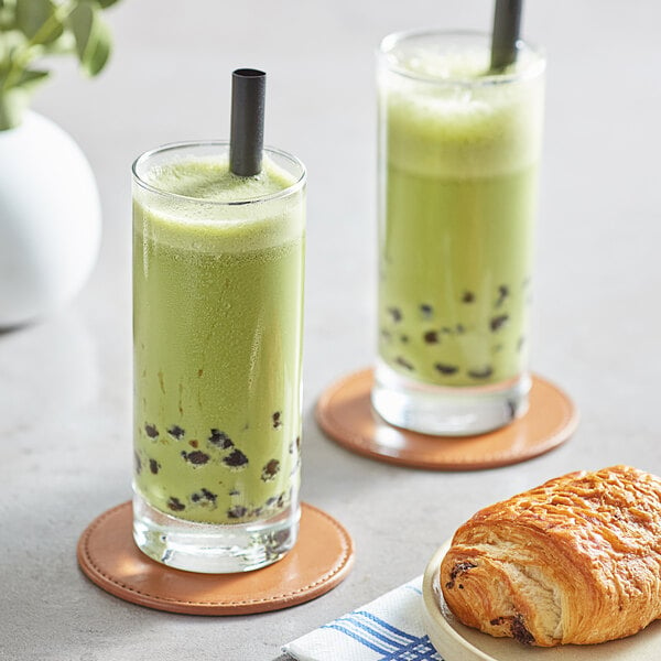 Two glasses of green matcha bubble tea with black straws and a croissant.