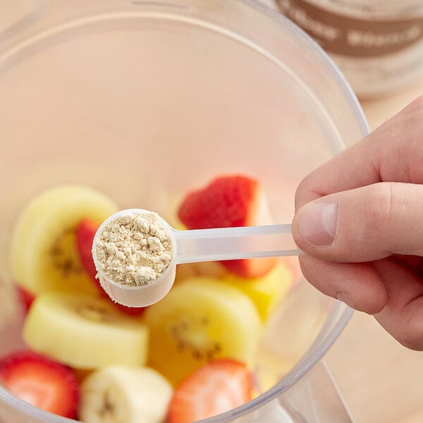 A hand holds a measuring spoon with Add A Scoop Fiber Blend supplement powder over a bowl of fruit.