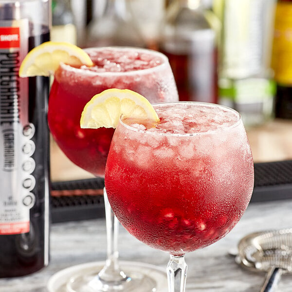 Two glasses of red liquid with lemon slices on a table with a bottle of SHOTT Pomegranate Real Fruit Flavoring Syrup.