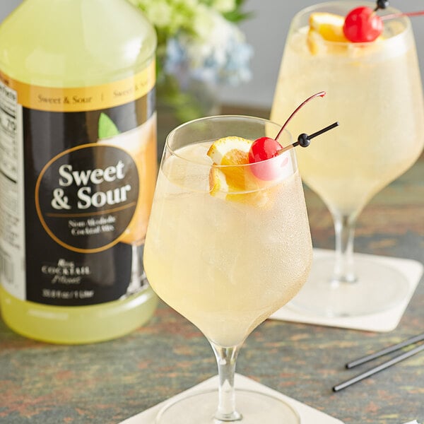 Two glasses of yellow liquid with a cherry and a lemon on top next to a bottle of Regal Cocktail Sweet and Sour Mix.