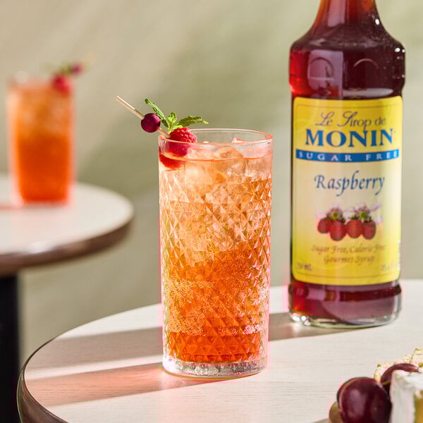 A bottle of Monin Sugar-Free Raspberry Flavoring Syrup next to a glass of iced raspberry beverage garnished with berries and mint.
