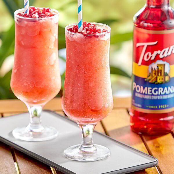 Two glasses of red drinks with straws and a bottle of red Torani Pomegranate flavoring syrup.