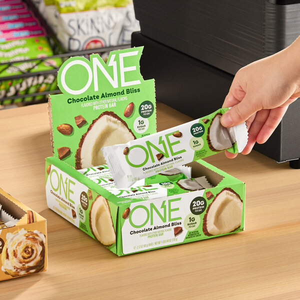A box of ONE Chocolate Almond Bliss protein bars with a hand picking up one bar.