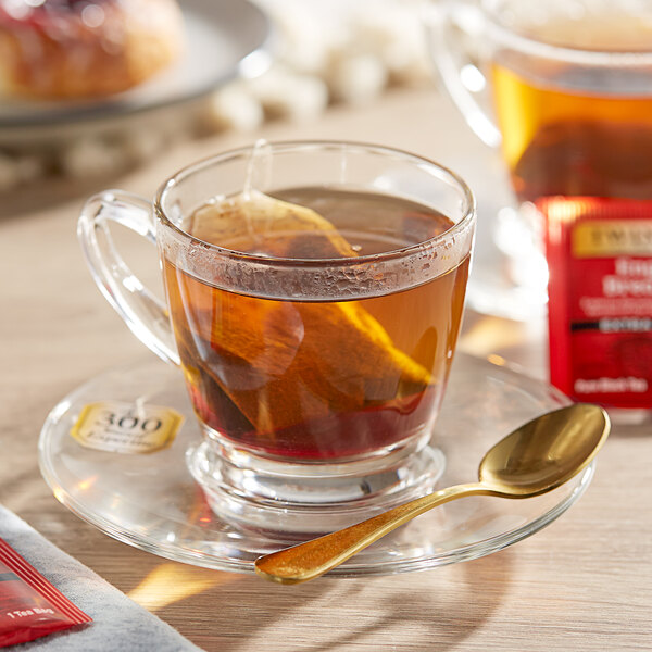 A glass cup of Twinings English Breakfast Extra Strong Tea with a tea bag and a spoon on a saucer.