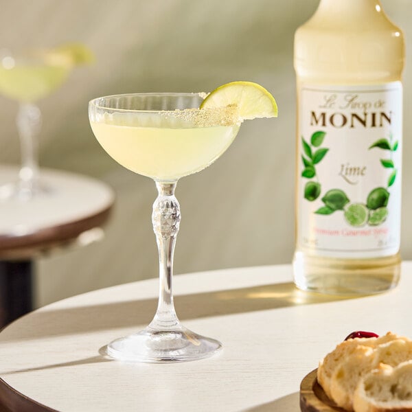A glass of lime-flavored cocktail garnished with a lime wedge, next to a bottle of Monin Premium Lime Syrup.