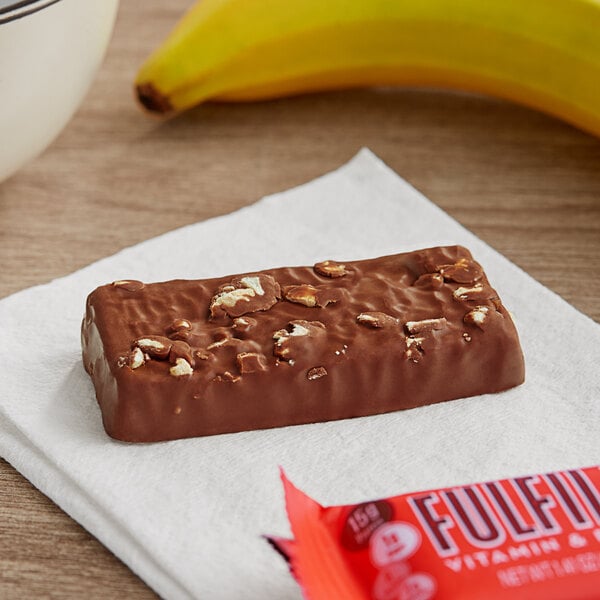 A red package of FULFIL chocolate peanut butter protein bars on a table with a banana.