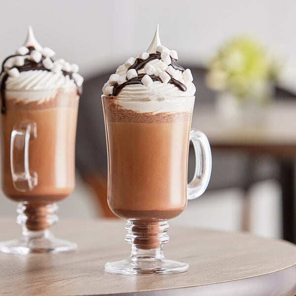 Two glasses of Capora hot chocolate with whipped cream and marshmallows on a table.