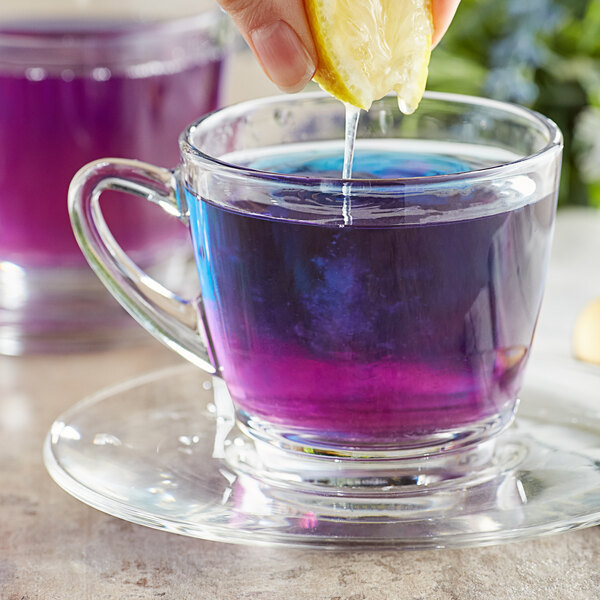 A person's hand squeezing a lemon over a glass cup of Wild Hibiscus Butterfly Pea Flower Tea