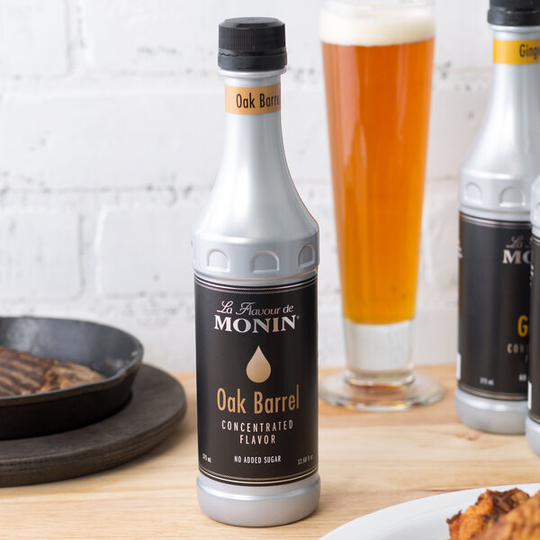 A black Monin Oak Barrel Concentrated Flavor bottle with gold text on a table next to a plate of food.