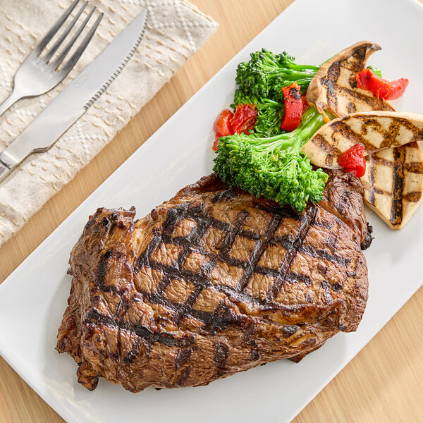 A grilled steak served with broccolini, grilled vegetables, and red peppers on a white rectangular plate.