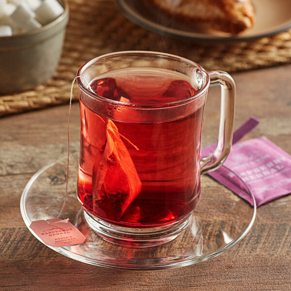 A glass mug of red Harney & Sons raspberry tea with a tea bag in it on a saucer.