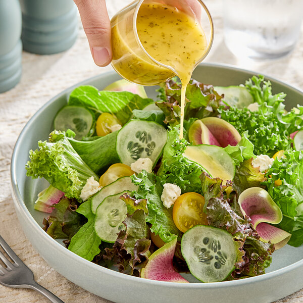 A fresh green salad with sliced cucumbers, cherry tomatoes, radishes, and crumbled cheese being drizzled with a yellow vinaigrette dressing.