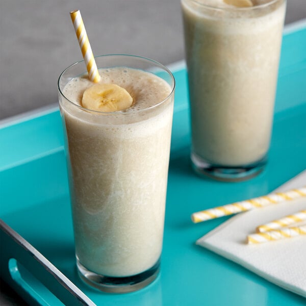 Two glasses of milkshake with DaVinci Gourmet Banana syrup, banana slices, and straws on a blue tray.