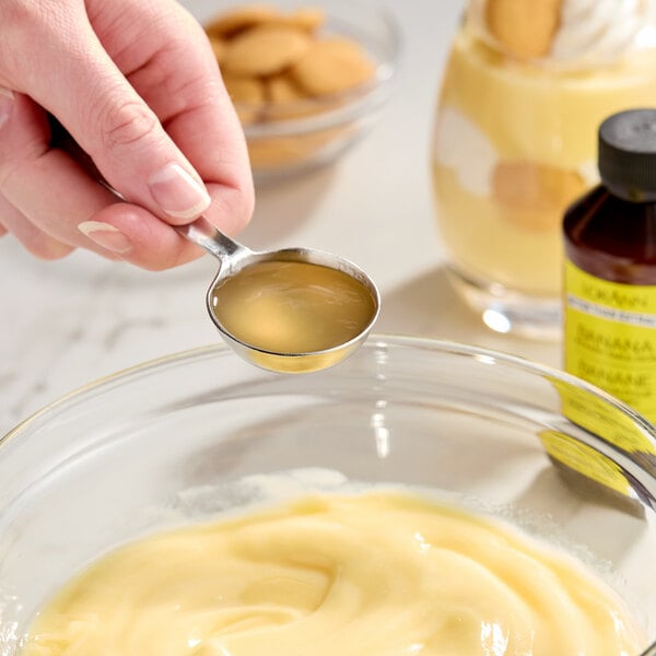 A hand holding a measuring spoon of liquid over a bowl of pudding, with a bottle of banana bakery emulsion and dessert items in the background.