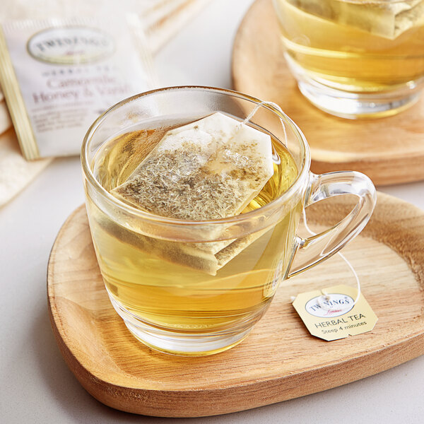 A glass mug and a glass cup of Twinings chamomile, honey & vanilla tea with tea bags in them on a wooden tray.