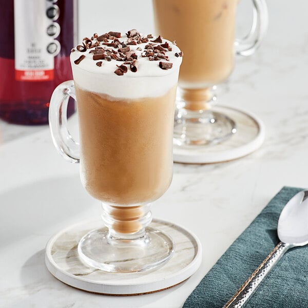 A glass mug of coffee with SHOTT Irish Cream syrup and whipped cream on a table with a spoon.