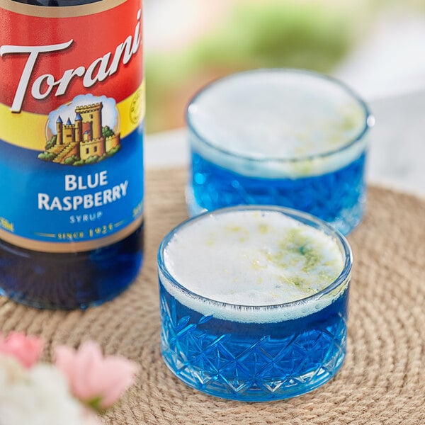 A glass of blue Torani raspberry flavoring next to a bottle of Torani Blue Raspberry syrup.