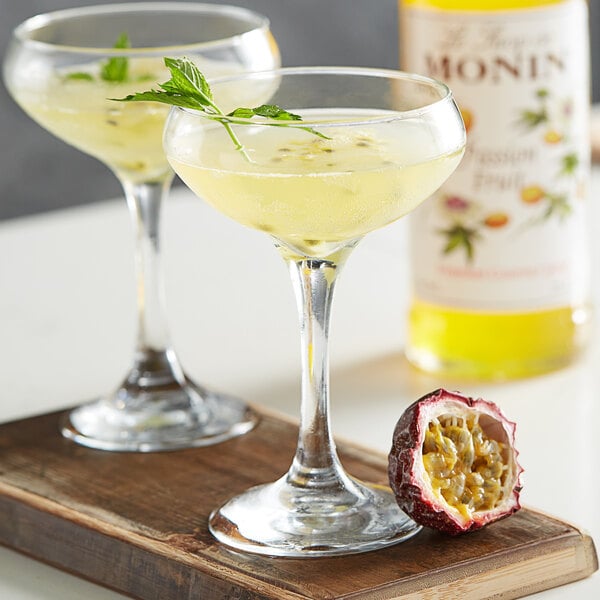 Two glasses of yellow liquid with Monin Passion Fruit Syrup and a half passion fruit on a table.