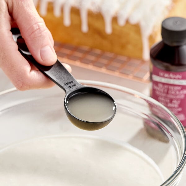 A hand holding a tablespoon of liquid over a glass mixing bowl, with a bottle of LorAnn Oils Buttery Sweet Dough Bakery Emulsion and a glazed loaf cake in the background.