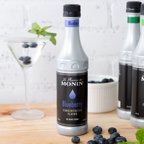 A bottle of Monin blueberry concentrated flavor on a table next to a glass of blueberries.