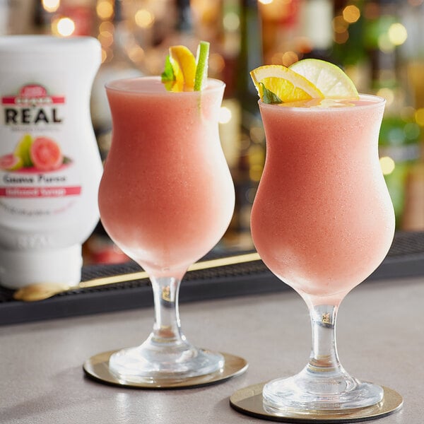 Two glasses of pink drinks garnished with lime on a table with a bottle of Real Guava Puree Infused Syrup.