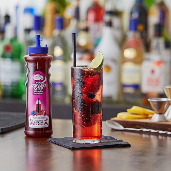 A glass of red liquid with a lime slice next to a bottle of Master of Mixes Raspberry Syrup.