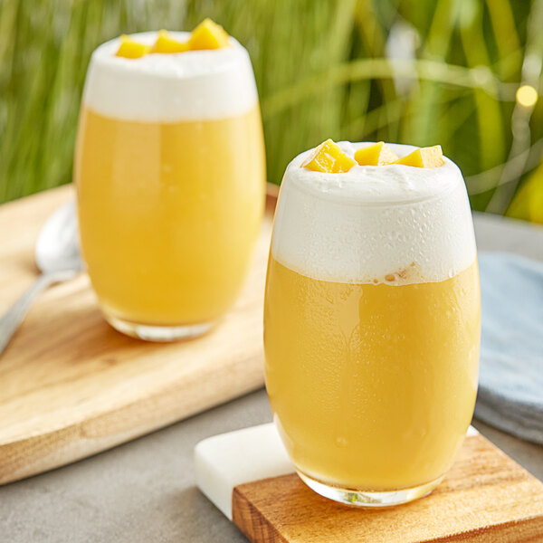 Two glasses of yellow mango drinks with white foam on top.