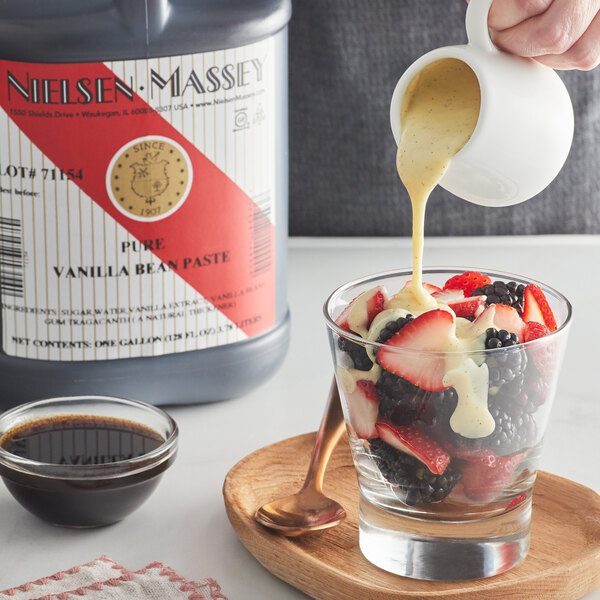 A person pouring Nielsen-Massey vanilla paste into a bowl of fruit with cream.