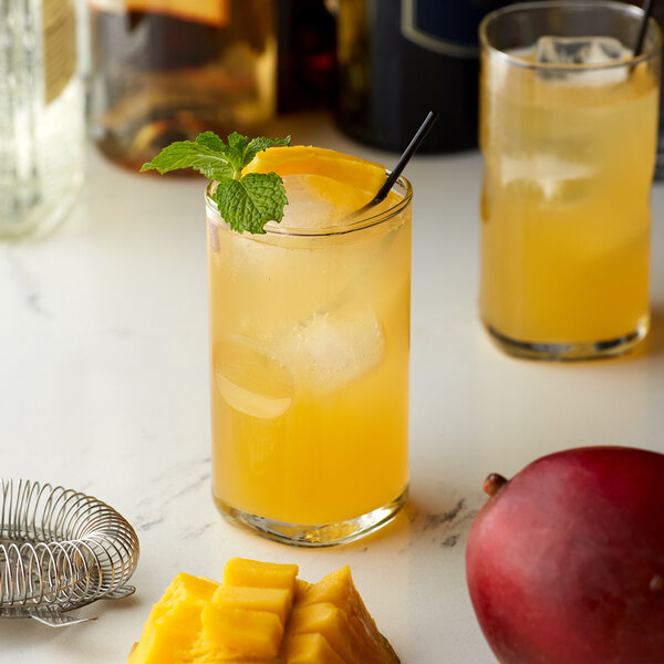 A glass of yellow liquid with a straw and a Monin Sugar Free Mango Flavoring Syrup with a mango slice on the rim.