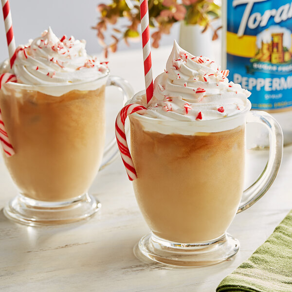 Two glass mugs of coffee with whipped cream and candy canes with a bottle of Torani Sugar-Free Peppermint Flavoring Syrup on the side.