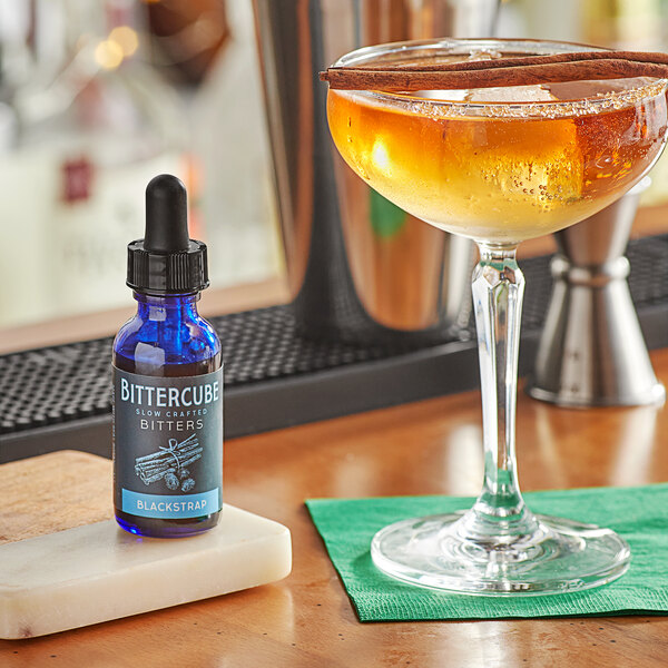 A glass of liquid with ice and a cinnamon stick next to a bottle of Bittercube Blackstrap Bitters on a table.