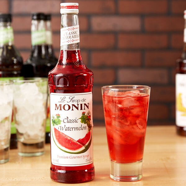 A bottle of Monin watermelon syrup next to a glass of watermelon soda with ice.