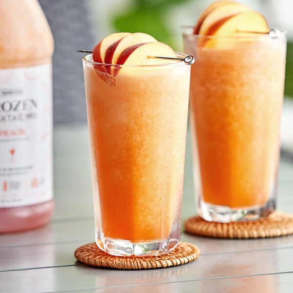 Two glasses of Narvon Peach frozen cocktail with peach slices on a table.