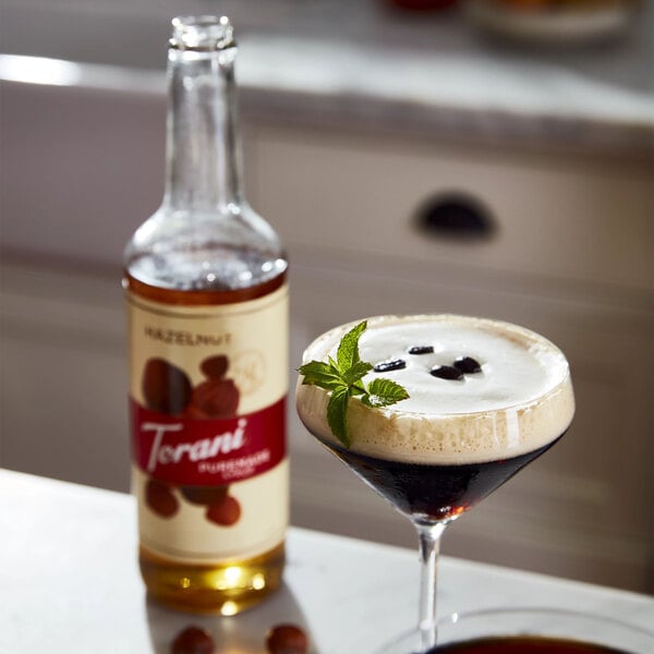 A glass of hazelnut-flavored liquid next to a bottle of Torani Puremade Hazelnut Flavoring Syrup.