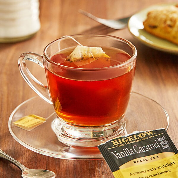 A package of Bigelow Vanilla Caramel Tea bags on a table with a glass cup of tea with a tea bag on a saucer.