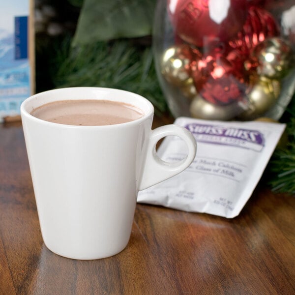 A white mug of Swiss Miss No Sugar Added hot chocolate on a table next to a Christmas tree.