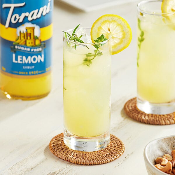 A pair of glasses of lemonade with lemon slices and herbs on a table with a bottle of Torani Sugar-Free Lemon Flavoring.