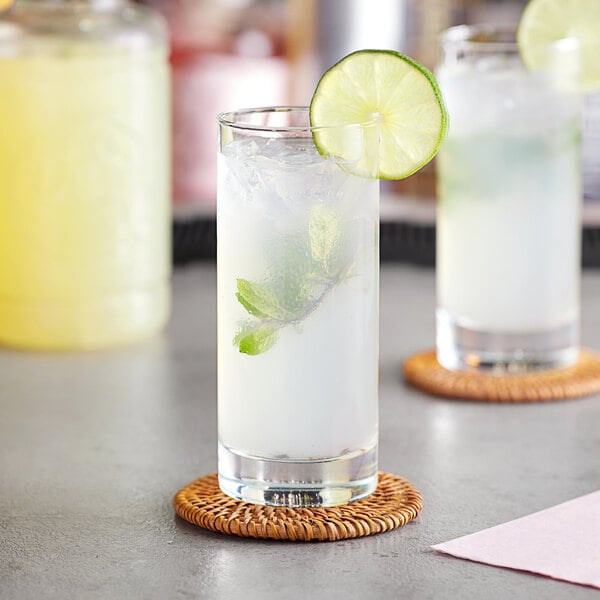 A glass of yellow liquid with ice and a lime slice with a leaf in it.