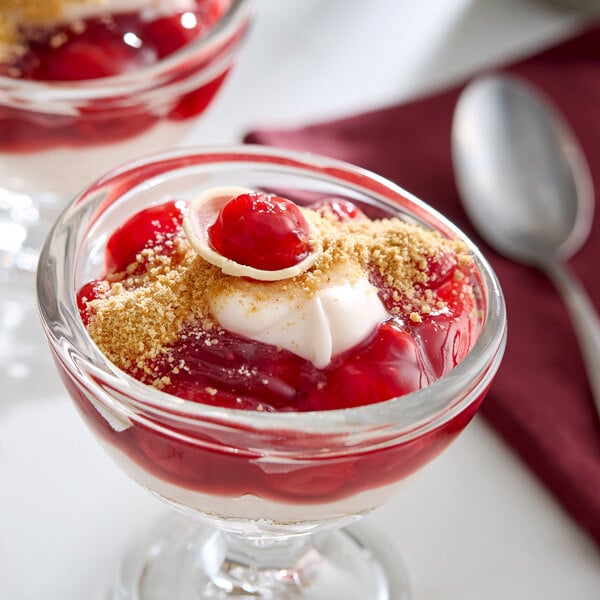 A glass dessert dish filled with cherry topping, whipped cream, and graham cracker crumbs.